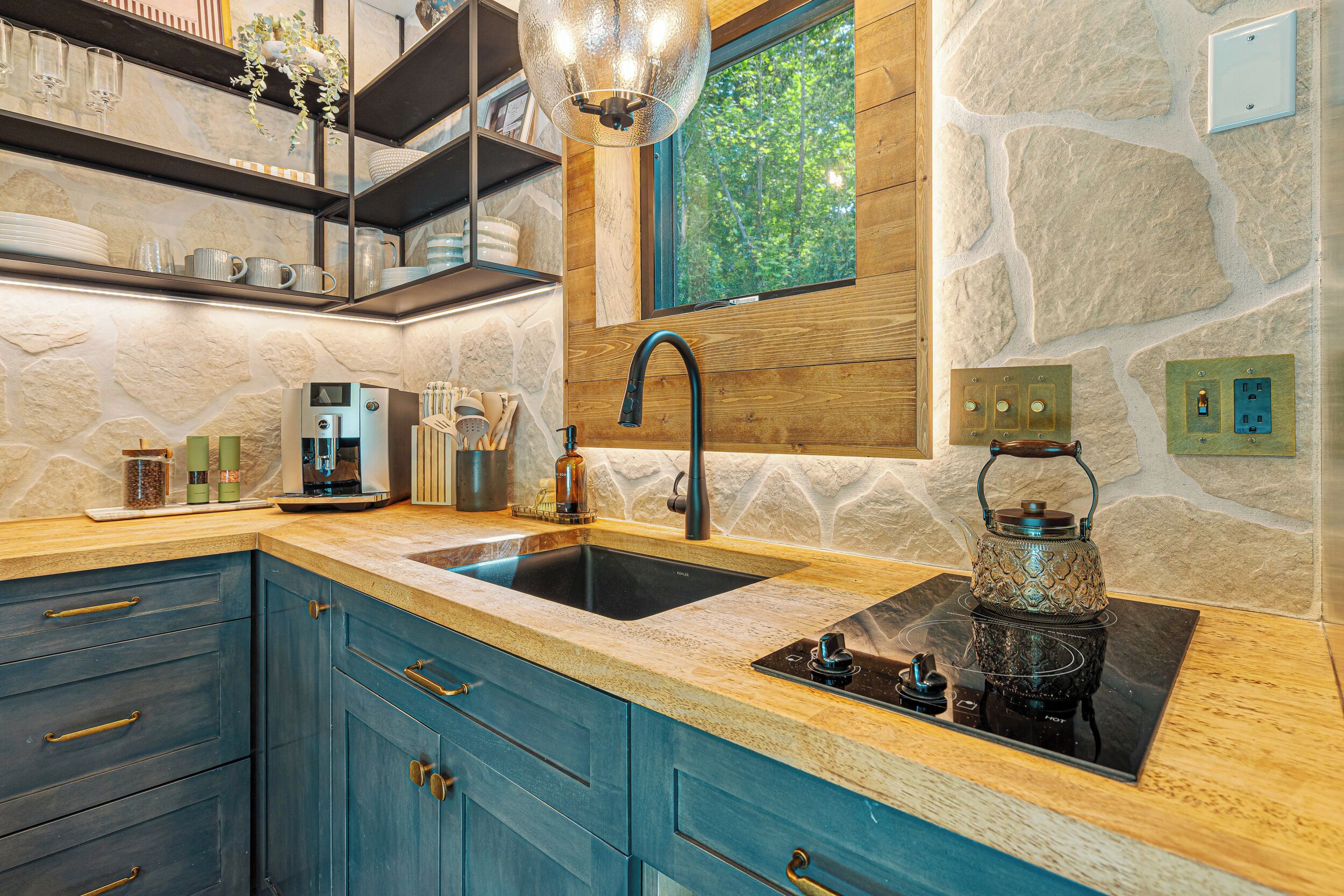 A modern kitchen with a stone wall backsplash, a built-in sink, and a window overlooking greenery.