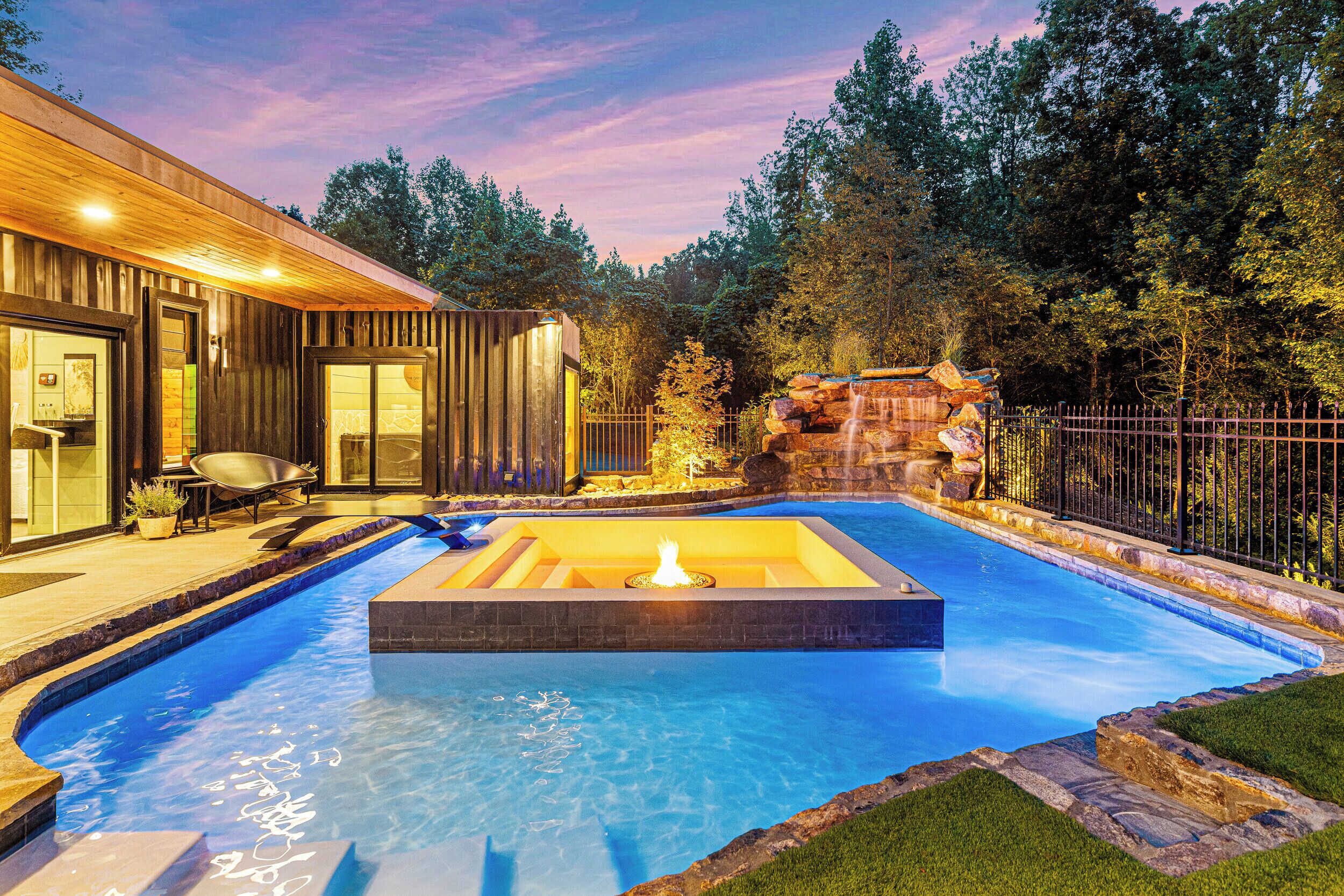 A swimming pool with a hot tub, surrounded by a wooden deck and a stone waterfall feature.