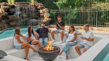 A group of people sitting around a fire pit by a pool with a waterfall in the background.