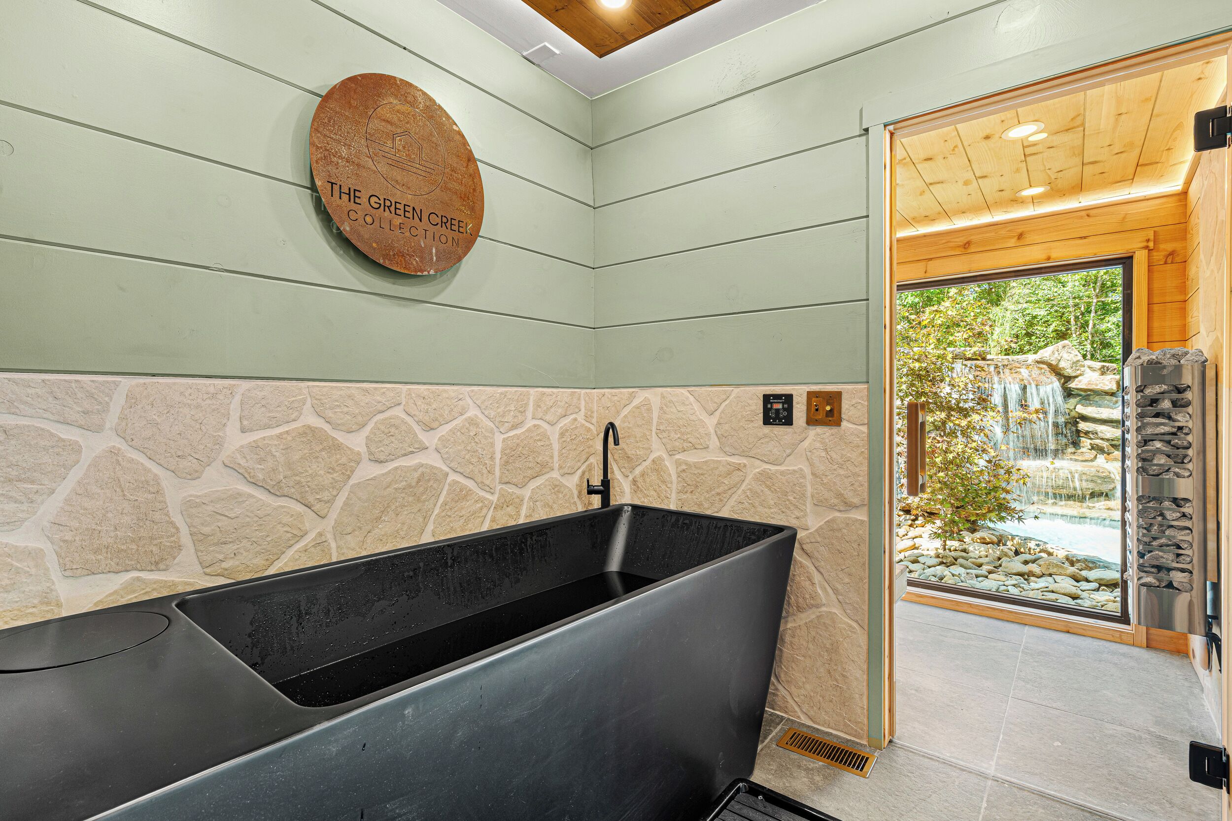 A bathroom with a large black bathtub, stone wall backsplash, and a wooden door leading to a natural outdoor area.