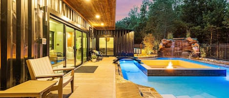 A modern outdoor pool area with a wooden deck, lounge chairs, and a stone waterfall feature.