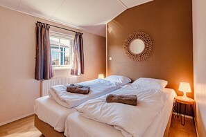Quarto de hotel com duas camas, uma janela com cortinas, um espelho fixado na parede e uma mesa de cabeceira com abajur.