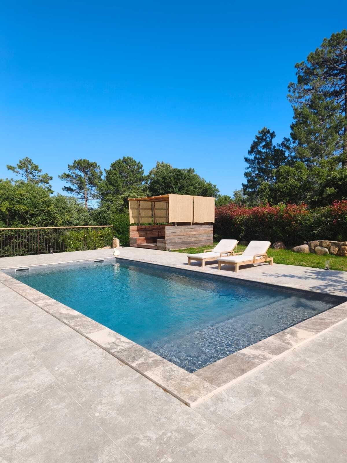 A rectangular swimming pool with a stone deck, two lounge chairs, and a wooden structure with a canopy.
