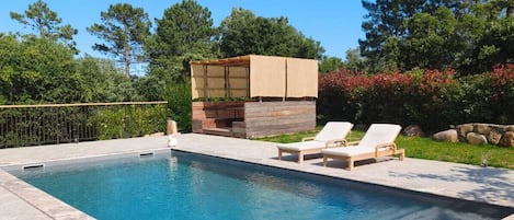 A rectangular swimming pool with a stone deck, two lounge chairs, and a wooden structure with a canopy.