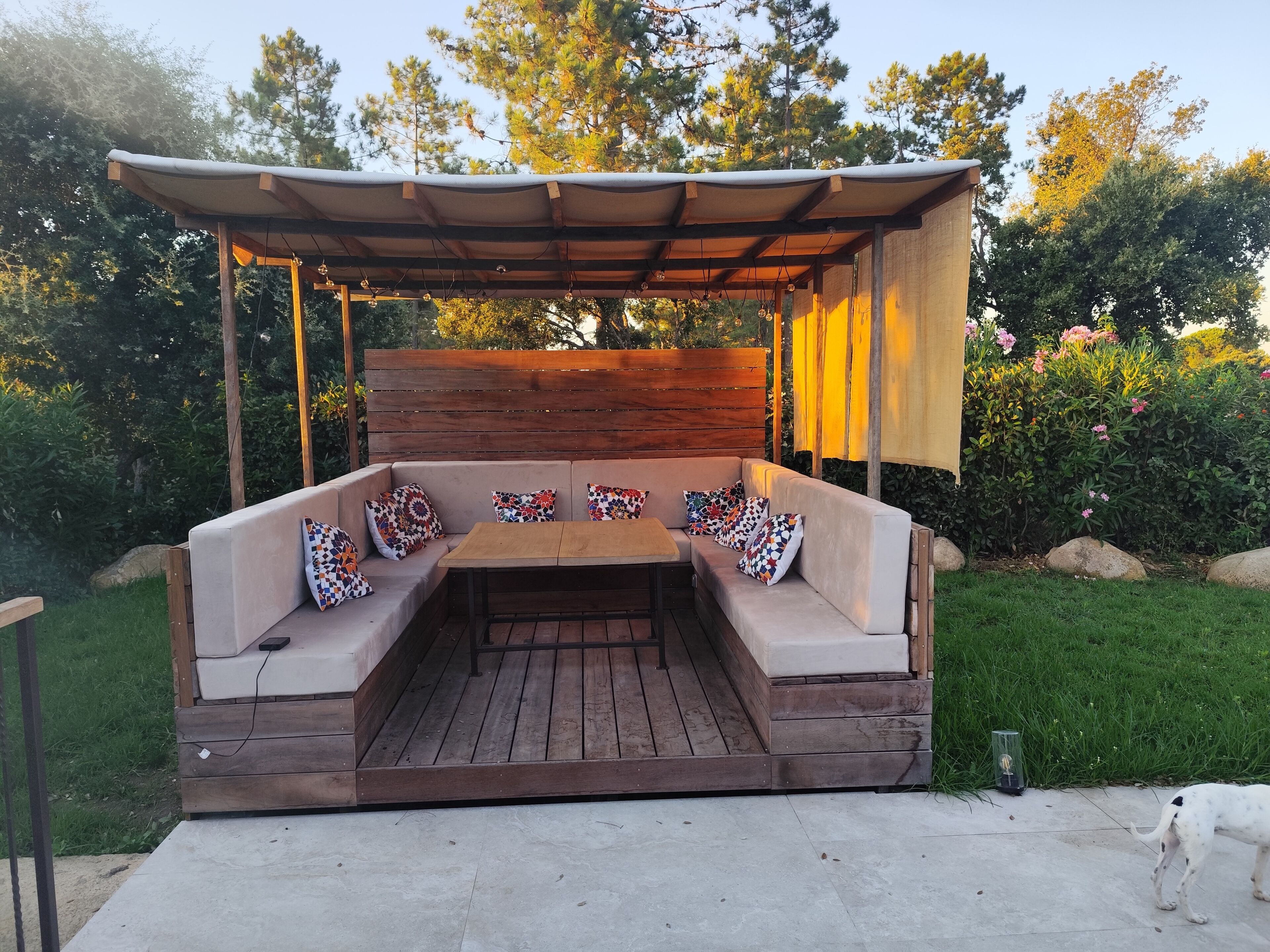 Een terras met een houten bank en kussens, omgeven door groen en een houten hek.