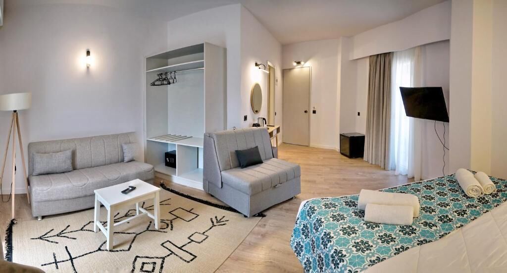 Una habitación de hotel moderna con sofá, una mesita, una silla, una cama con colcha estampada, televisor y armario.
