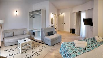 Una habitación de hotel moderna con sofá, una mesita, una silla, una cama con colcha estampada, televisor y armario.