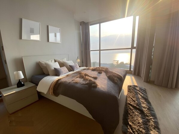 Un dormitorio moderno con una cama grande, una mesita de noche, una lámpara y vistas de la ciudad a través de amplias ventanas.