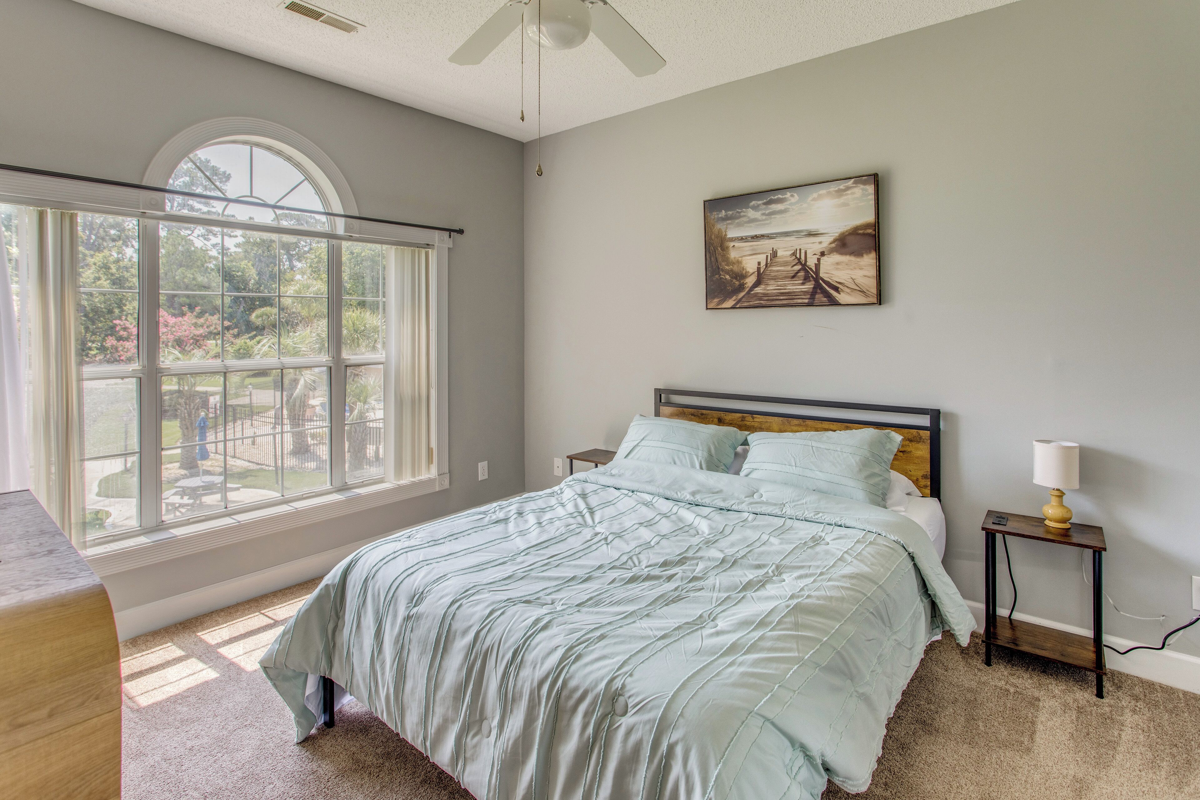 A bedroom with a large window, a bed with a headboard, a bedside table with a lamp, and a painting on the wall.