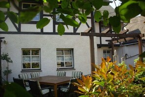 Eine Terrasse mit Holztisch und -stühlen, ein weißes Gebäude mit schwarzer Verzierung und eine Pergola.