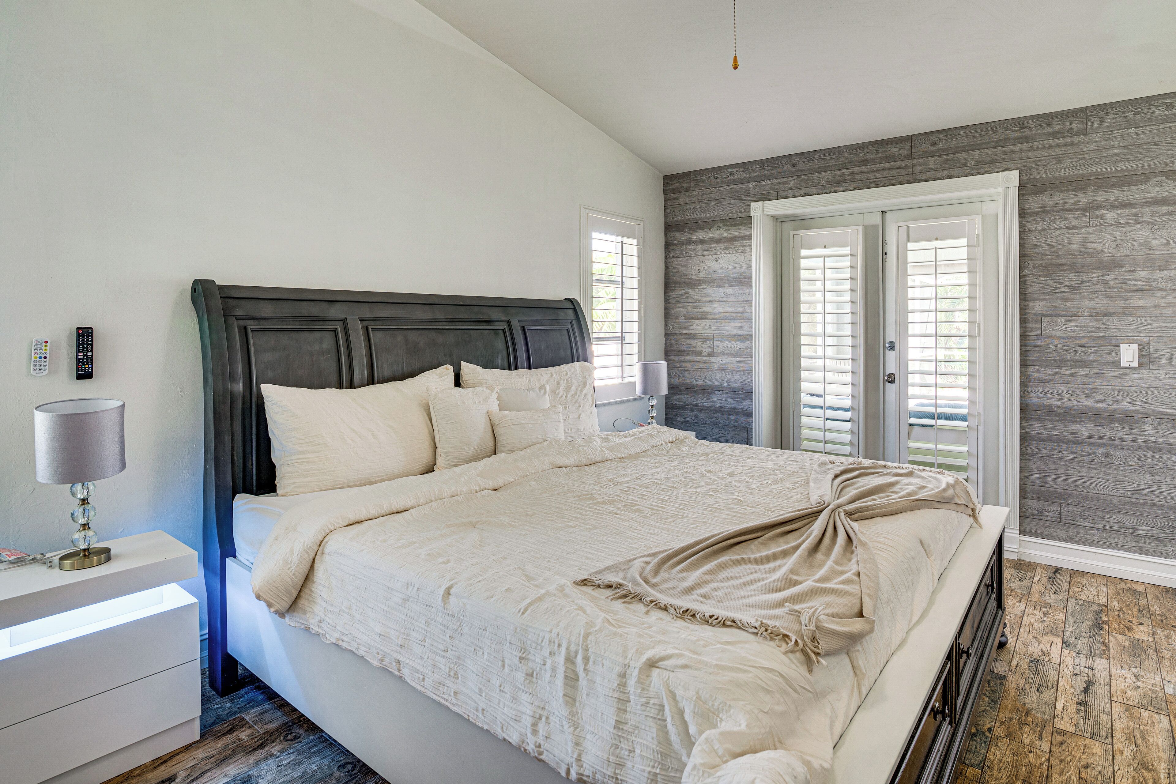 A bedroom with a large bed, bedside table, lamp, and a view of the outside through a door.