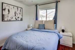 A bedroom with a large bed, a bedside table with a lamp, a wall-mounted artwork, and a window with curtains.