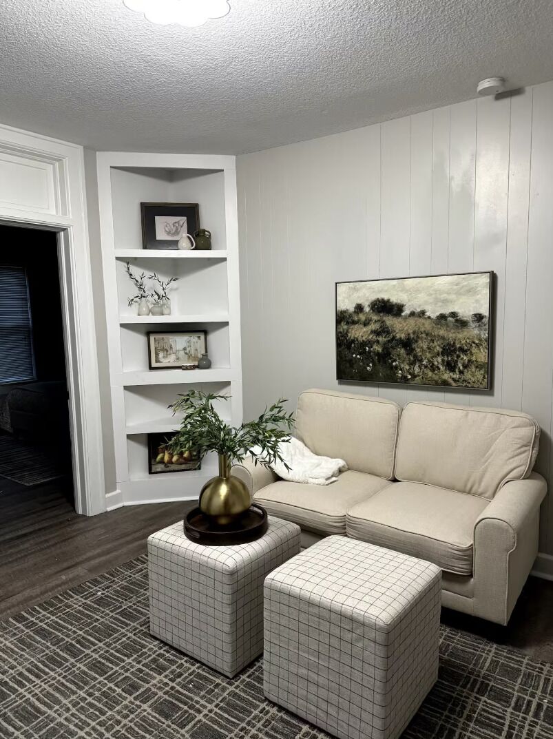 A living room with a beige sofa, a white wall with shelves, a framed painting, and a patterned rug.