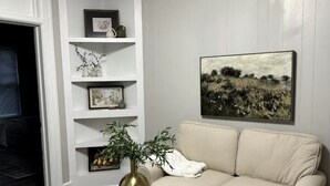 Ein Wohnzimmer mit einem beigen Sofa, einer weißen Wand mit Regalen, einem gerahmten Gemälde und einem gemusterten Teppich.