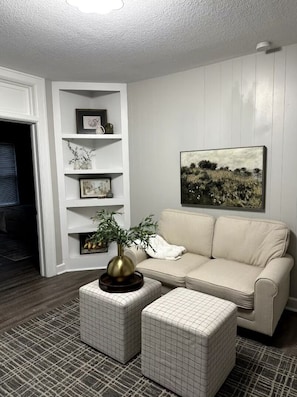 Ein Wohnzimmer mit einem beigen Sofa, einer weißen Wand mit Regalen, einem gerahmten Gemälde und einem gemusterten Teppich.