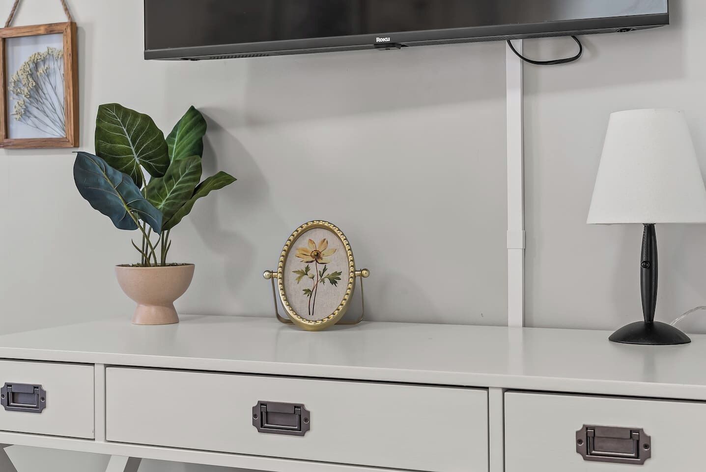A white dresser with drawers, a potted plant, a framed picture, a lamp, and a television mounted on the wall.