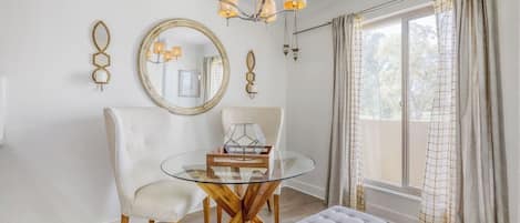 A room with a round glass table, two chairs, a bench, a chandelier, a large mirror, and a window with curtains.