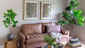 A living room with a brown leather sofa, two framed artworks on the wall, and two potted plants.
