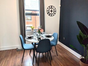 Ein Essbereich mit einem runden Tisch, gedeckt für vier Personen, zwei blauen Stühlen, einer Wanduhr und einem Fenster mit Vorhängen.
