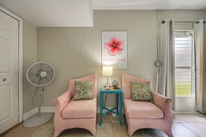 Ein Zimmer mit zwei rosafarbenen Sessel, einem türkisfarbenen Beistelltisch, einem Ventilator und einem Fenster mit Vorhang.