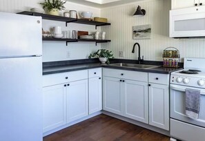 A kitchen with white cabinets, a black countertop, a sink, a stove, and shelves with kitchenware. - 5 Min to Parkview, Attached Garage & Office (Fort Wayne)