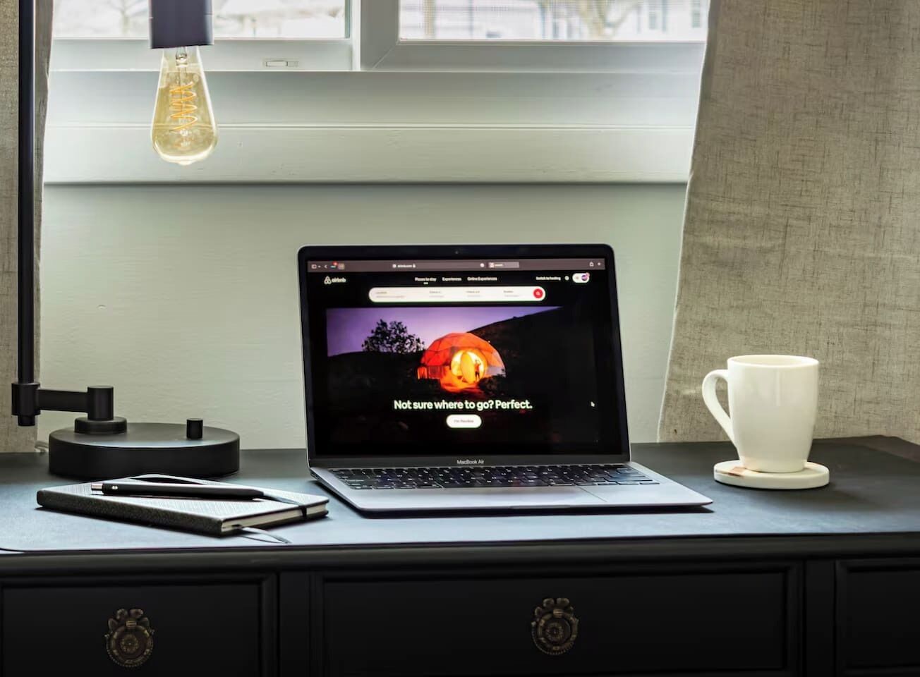 A laptop on a desk with a cup and a notebook beside it.