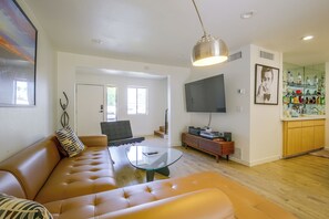 Ein modernes Wohnzimmer mit einer Ledersofa, einem Glastisch, einem Flachbildfernseher und einer Küchenzeile mit Holzschränken und einem Weinkühlschrank.