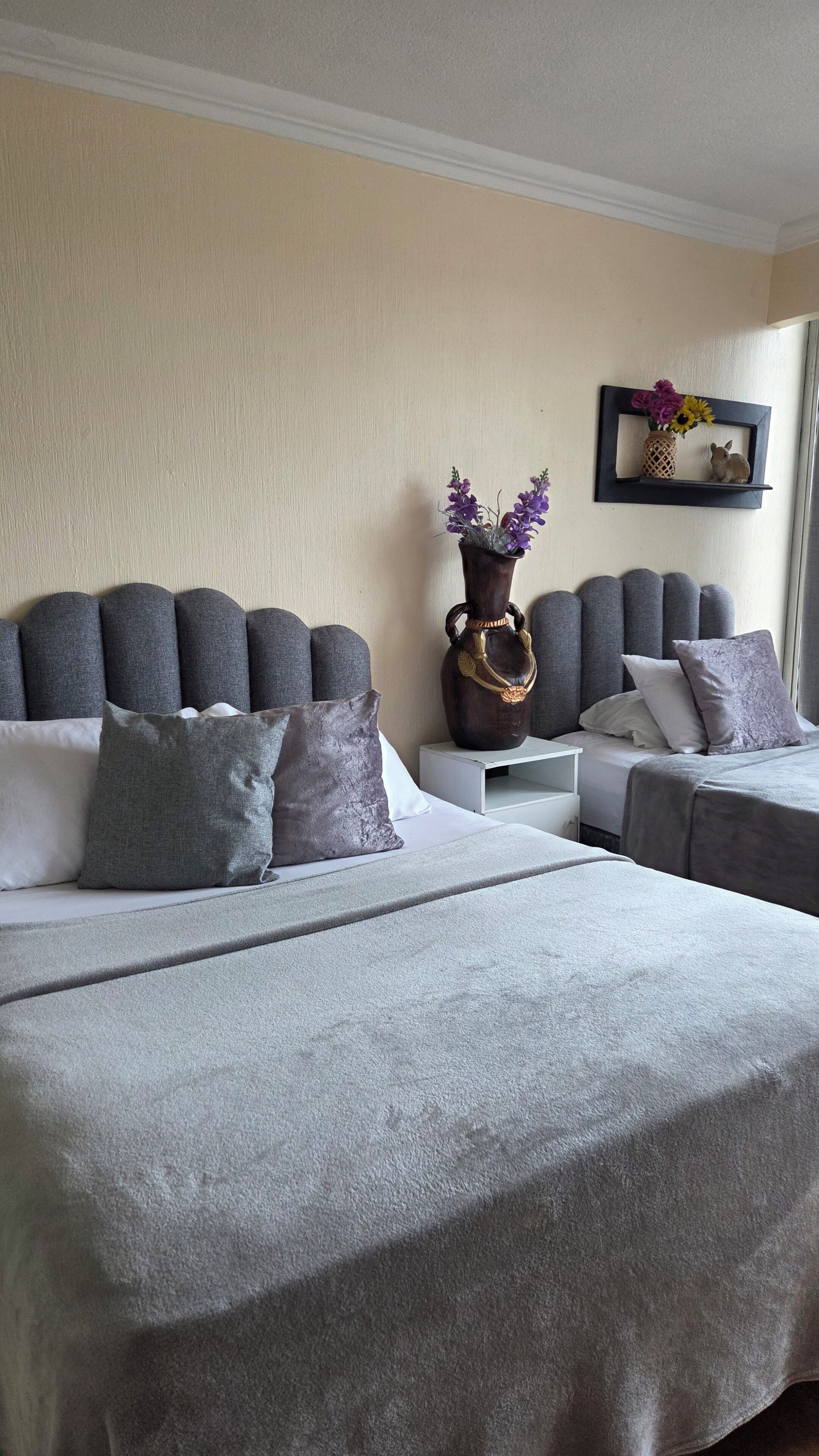 Une chambre d’hôtel avec deux lits, une literie grise, un vase décoratif avec des fleurs et une petite étagère sur laquelle est posé un cadre photo.