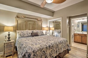 A bedroom with a large bed, two bedside lamps, a wooden nightstand, a headboard with decorative items, and a bathroom visible through an open door. - Gulf Views & Beach Access! 2 PCB Condos w/ Pool! (Panama City Beach)