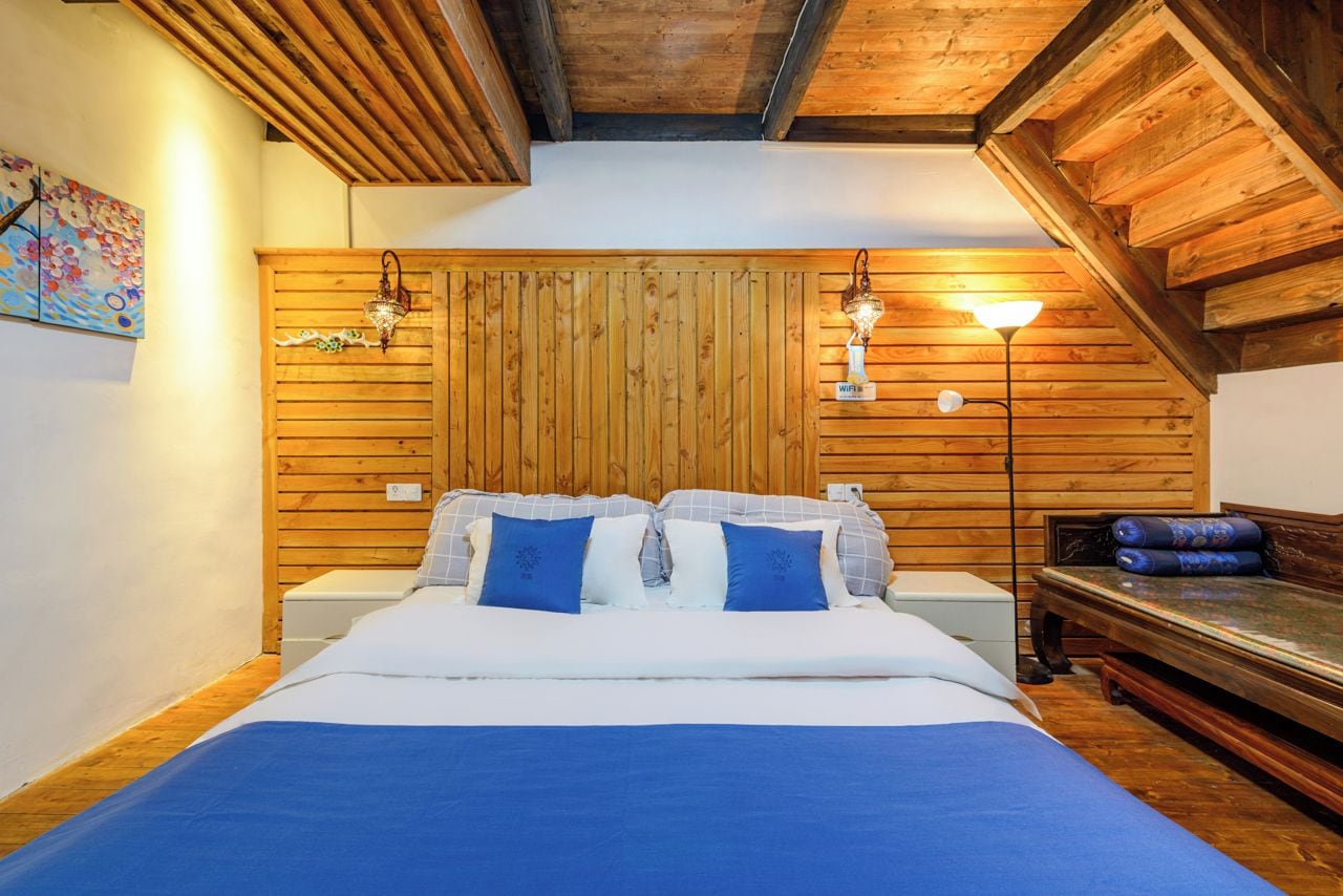 A bedroom with a wooden slatted headboard, a bed with blue bedding, two bedside lamps, and a wooden bench with cushions.