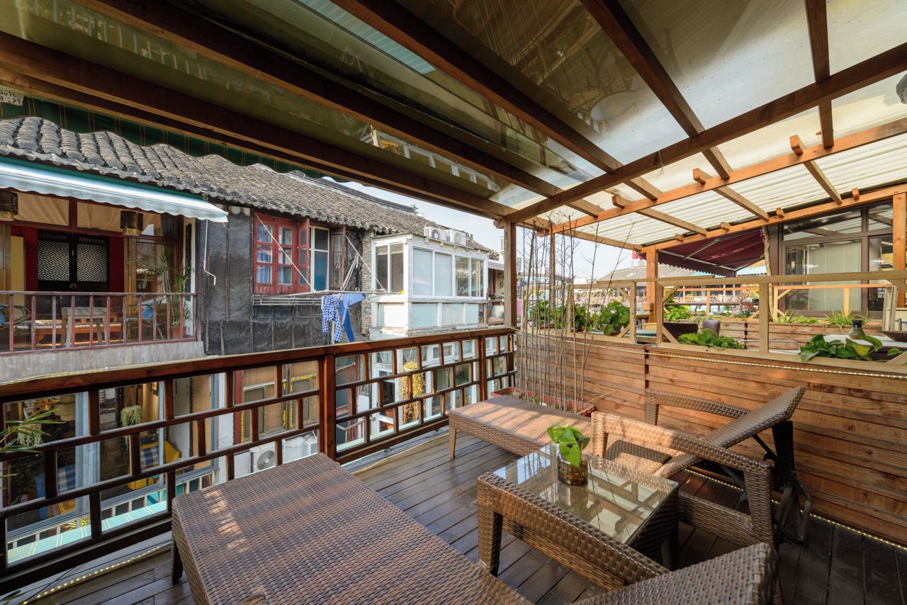 Una terraza de madera tradicional con mobiliario de mimbre y una mesa de vidrio, con vista a un patio con plantas y un edificio con tejado de baldosas.