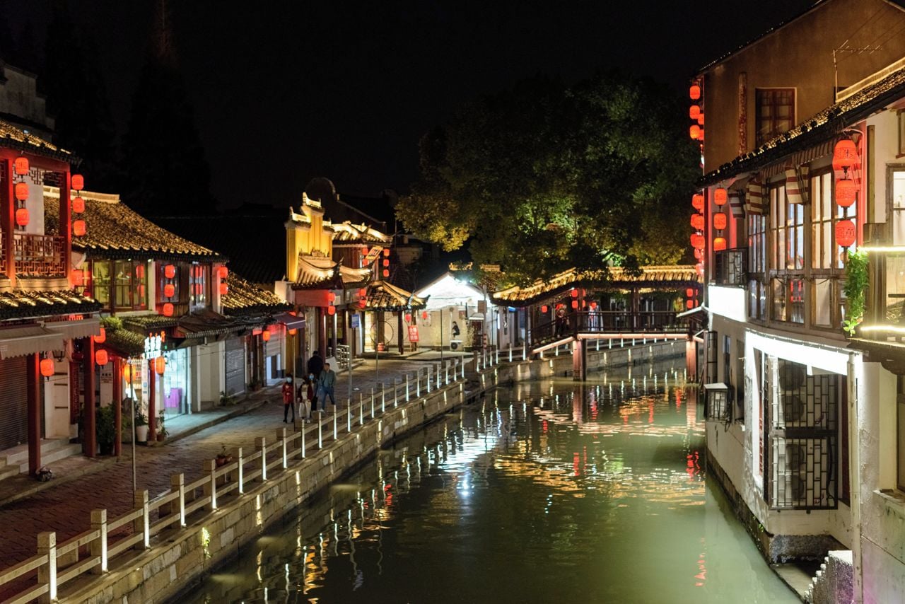 A narrow waterway flanked by traditional buildings with red lanterns, a wooden bridge, and a clear night sky.