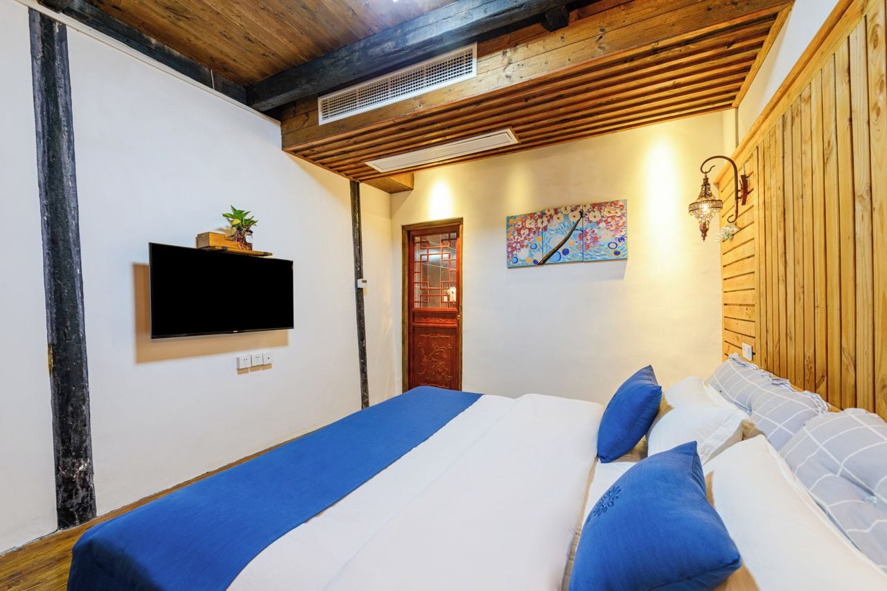 A bedroom with a bed, a flat-screen TV, a wooden ceiling, a door, and a wall-mounted light fixture.