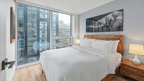 Quarto de hotel moderno com uma cama grande, cabeceira de madeira, duas mesas de cabeceira com abajures, uma janela ampla com vista para edifícios e uma obra de arte emoldurada na parede.
