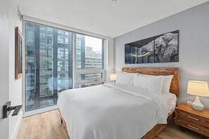 A modern hotel room with a large bed, a wooden headboard, two bedside tables with lamps, a large window with a view of buildings, and a framed artwork on the wall. - 1 Bedroom/1 Bathroom Suite (Montreal)