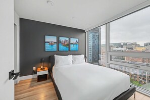 A modern bedroom with a large bed, a nightstand, a wall-mounted TV, and a city view through a large window. - 2 Bedroom/2 Bathroom Suite (Montreal)