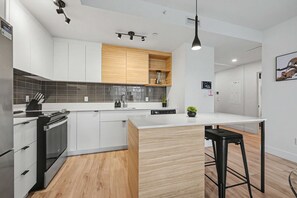 A modern kitchen with white cabinetry, a wooden accent wall, a built-in island, and a bar stool. - 2 Bedroom/2 Bathroom Suite (Montreal)