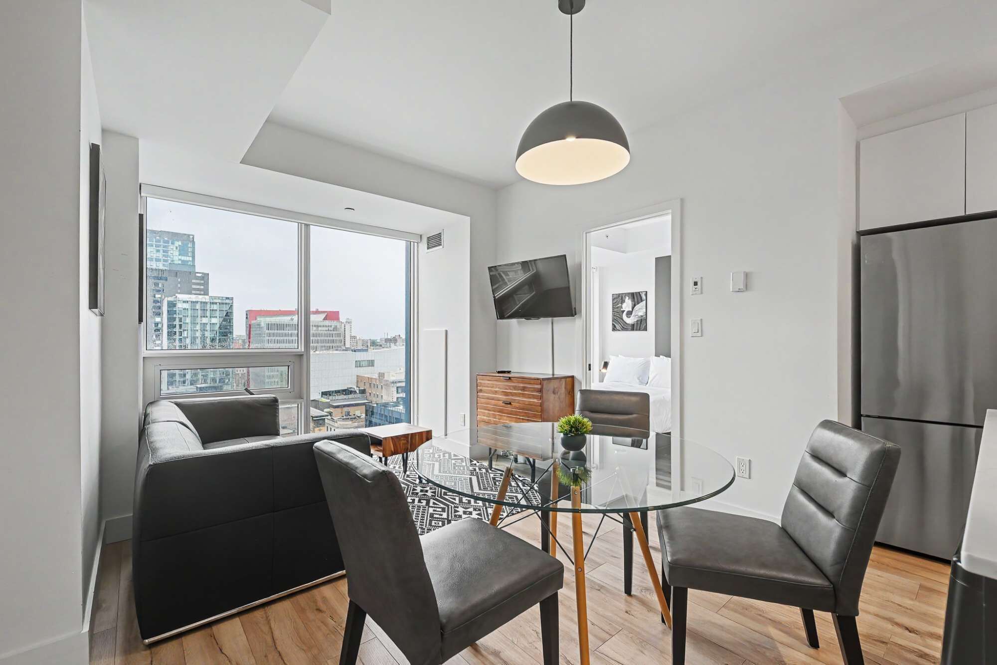 Una cocina moderna con refrigerador, un comedor con mesa y sillas de vidrio, y vista a la ciudad a través de amplios ventanales.