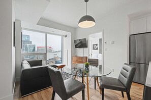 A modern kitchen with a refrigerator, a dining area with a glass table and chairs, and a city view through large windows. - 2 Bedroom/2 Bathroom Suite (Montreal)