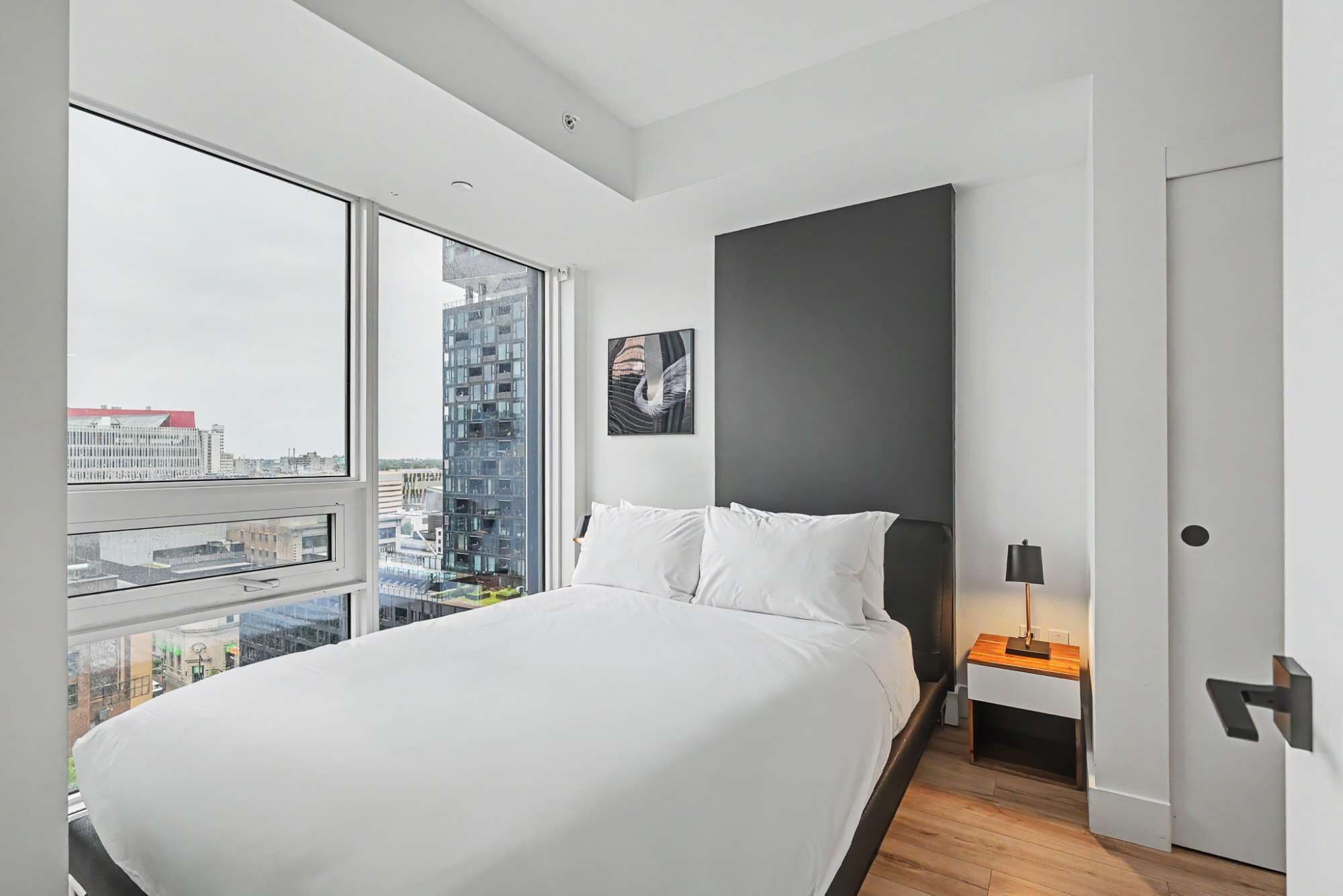 A modern bedroom with a large bed, a nightstand, a lamp, and a city view through the window.