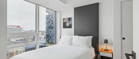 A modern bedroom with a large bed, a nightstand, a lamp, and a city view through the window.