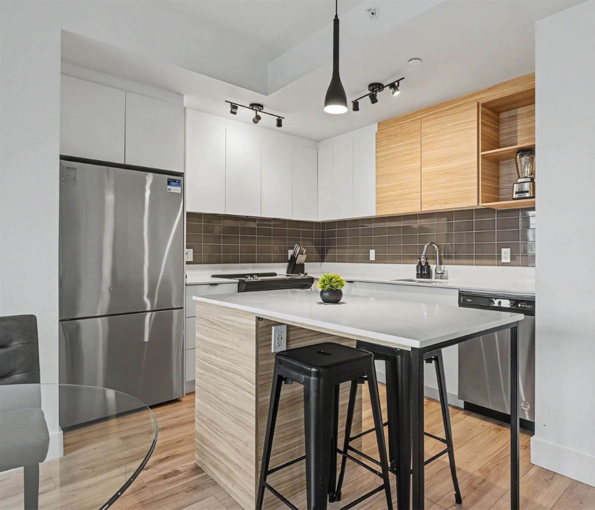 A modern kitchen with a stainless steel refrigerator, a central island with stools, a wooden upper cabinet, and a backsplash.
