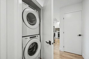 A laundry room with two stacked Whirlpool washers and dryers, a white door, and a hallway leading to a kitchen area. - 2 Bedroom/2 Bathroom Suite (Montreal)