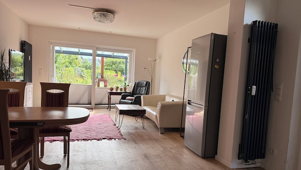 A modern living room with a dining area, a black refrigerator, and a view of greenery outside.