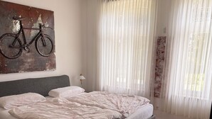 A bedroom with a bed, a wall-mounted bicycle art piece, sheer curtains, and a wooden floor.
