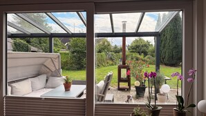 A room with a glass ceiling, a sofa, a table, and a view of a garden with potted plants and a dog.