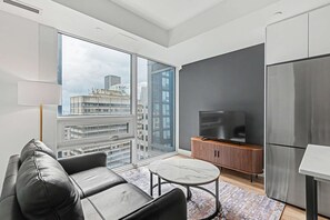 A modern living room with a sofa, a coffee table, a TV, and a large window offering a city view. - 1 Bedroom/1 Bathroom Suite (Montreal)