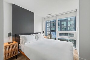 A bedroom with a bed, a nightstand, a lamp, and a large window with a city view. - 1 Bedroom/1 Bathroom Suite (Montreal)