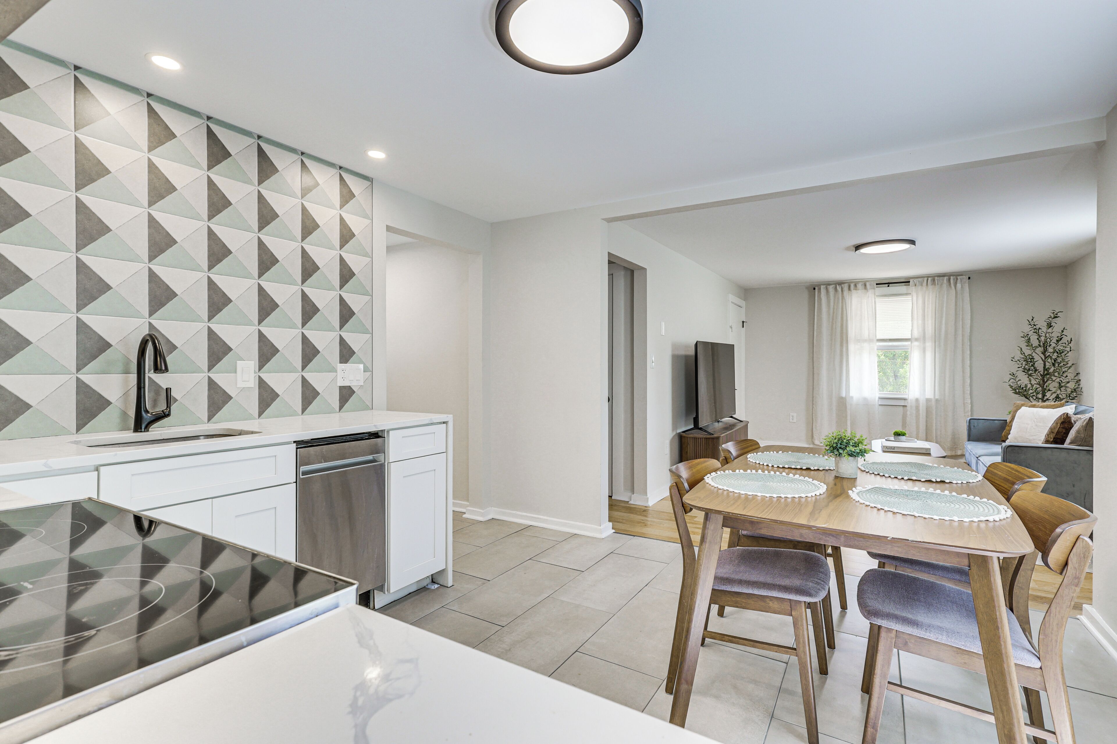 Une cuisine moderne avec un revêtement mural géométrique, un coin repas avec une table et des chaises en bois, et un salon avec un canapé et une plante.