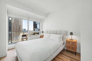A modern bedroom with a large bed, a nightstand, a chair, and a window with a city view. - 2 Bedroom/2 Bathroom Suite (Montreal)